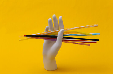 White mannequin hand holding many plastic cocktail tubes on yellow background