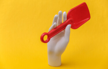 White mannequin hand holds red toy shovel on yellow background