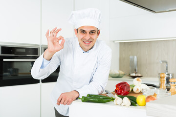 Young male cook in white uniform showing ok hand sign on kitchen