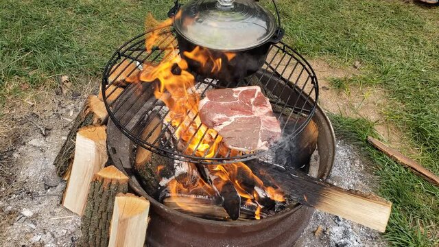 Placing t-bone beef steak and grilling over fire pit next to cost iron pot of boiling soup while at camp site.