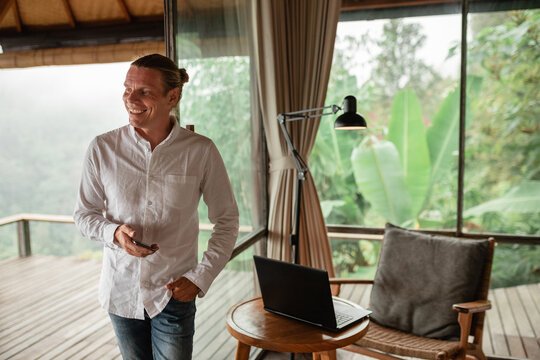 Young modern businessman hold in hands smart phone, smiling  while stand in the office near  working place with laptopp