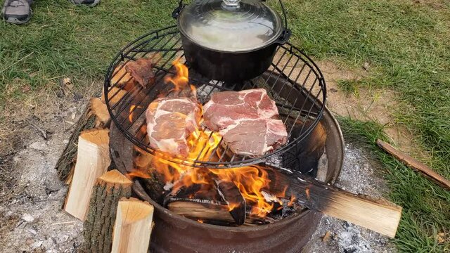 Placing beef steak and grilling over fire pit next to cost iron pot of boiling soup while at camp site.