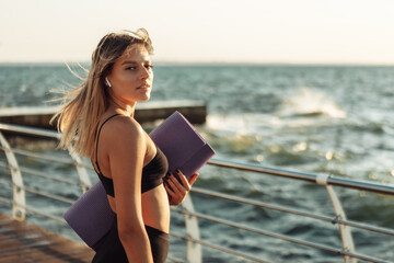 Young slim fit woman holding yoga mat at sunrise in the morning beach, before workout.