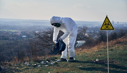 Full length of ecologist in protective suit and gas mask picking up garbage in abandoned grassy...