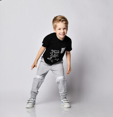 Frolic blond kid boy in sunglasses, black t-shirt with dinosaur print and gray pants stands leaning...