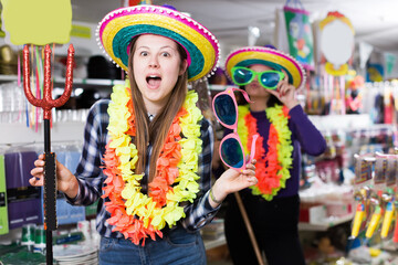 Comically dressed positive girl joking while visiting festive accessories shop with her female friend.