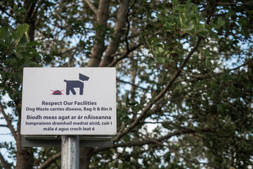 Sign with message in English and Irish language: Respect our facilities, dog waste carries disease, bag it, bin it. Sign mounted on a metal pole in a park.