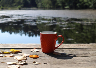 A mug of tea stands on the Bank of a bridge on a lake with yellow leaves, close - up-the concept of pleasant trips in autumn