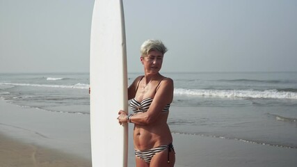 Portrait of mature active pensioner woman with surfboard on the sea beach