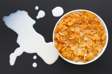 Corn flakes with milk in a white round bowl on a black metallic surface, and some milk spilled outside the bowl.