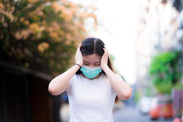 Beautiful woman held her head with both hands. Asian women wearing a green medical face mask stands in a public area. Concept of epidemic stress in which there is no cure and vaccine.