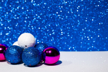Decorative Christmas balls in blue, white and purple on the left on a white surface. Blue Christmas lights on the background. Side view.Place for inscriptions and greetings