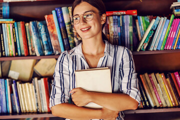 Young attractive female student in dress standing in library in front of shelves with books and...