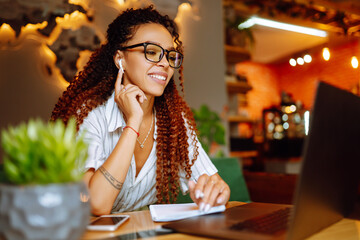 Portrait of African American woman sitting at cafe having video call on laptop computer. Woman, student wearing headphones calling on laptop, talk by webcam  with friend or relative, online course.