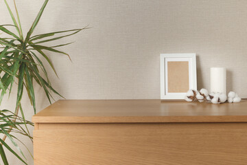 Wooden chest of drawers with a white candle, a white photo frame with a cotton branch on a light wall, on the left is a living plant dracaena - obrazy, fototapety, plakaty