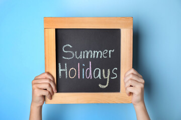 Schoolgirl holding blackboard with text SUMMER HOLIDAYS on light blue background, closeup