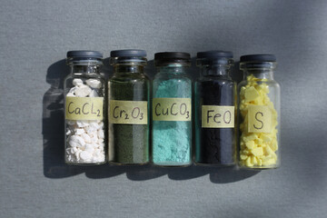 Large white crystals of calcium chloride, dark green chromium oxide, blue copper carbonate, black iron oxide, yellow large crystals of sulfur, in glass jars with the formulas.