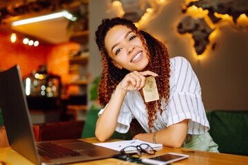 Young woman holding credit card and using laptop computer. African American woman sitting at cafe making online shopping. Online shopping, e-commerce, internet banking, spending money.