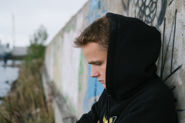 portrait of a sad lonely teenager / young guy in a black hoodie in an abandoned place near the port