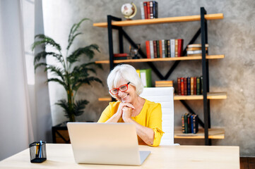 A touched gray-haired business lady in eyeglasses is looking at the laptop screen while sitting in a smart office
