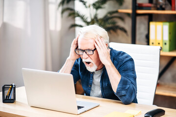 Senior man looks at the laptop screen with shock and surprise, he holding head in his hands