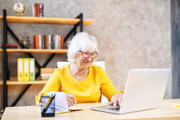 Older woman in smart casual wear and eyeglasses using laptop for work indoor. Beautiful grey-haired lady in the office