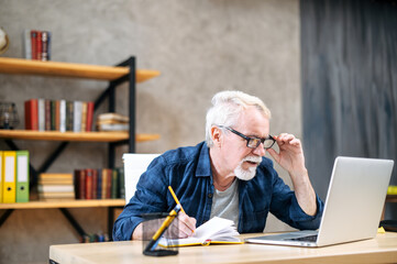 Focused older man in smart casual wear is using laptop computer for watching online classes and webinars, he is takes a notes at notebook, while sitting at contemporary home office