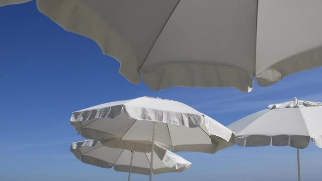 Fluttering frills of white textile umbrellas against blue sky background with copy space. Beach parasols on beach. Seaside relaxation concept