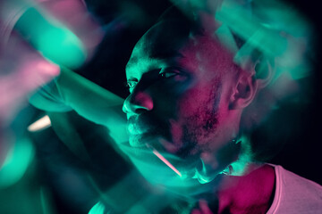 Evening mood. Cinematic portrait of stylish young man in neon lighted room. Bright neoned colors. African-american model, musician indoors. Youth culture in party, festival style and music concept.