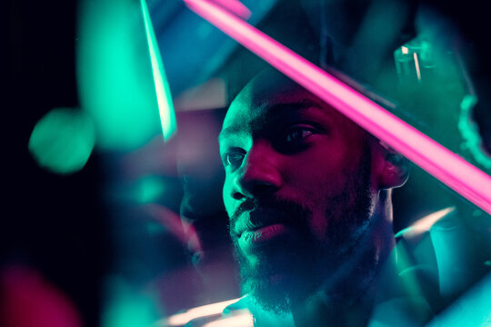 Evening mood. Cinematic portrait of stylish young man in neon lighted room. Bright neoned colors. African-american model, musician indoors. Youth culture in party, festival style and music concept.
