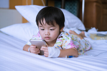 Asian cute little boy is looking smartphone and lying on the sickbed with saline intravenous on hand in the hospital. Healthcare medical and people concept.