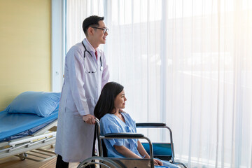 Fototapeta premium Young male doctor encouraging his female patient in hospital while pushing her wheelchair. Doctor informs report the illness while the patient is in wheelchair. Doctor looking after his patient.