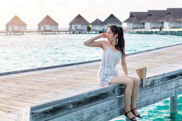 Beautiful young woman sitting on wooden pier by beautiful summer sun. Young Woman in white on a wooden beach pier. Girl enjoying vacation on wooden pier with water bungalow villa. Travel and Vacation.