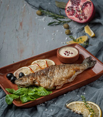 food photography, fried fish close-up side view, served with lemon, herbs and spices on a ceramic brown dish, against a gray textured background