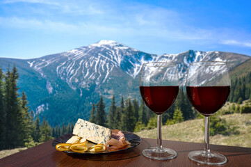 Two glasses of wine with cheese and meat assortment on view of mountains landscape. Glass of red wine with different snacks - plate with ham, sliced, blue cheese. Romantic celebration.