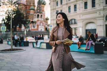 Sincerely female traveller with orientation paper map and smartphone gadget in hand exploring red square in Moscow during touristic journey in Russia, joyful woman in earbuds listening audio guide