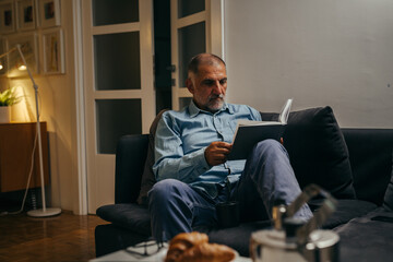 middle aged man sitting sofa reading book at home