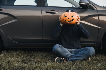 An unrecognizable adult man sits near a car on the grass and holds in front of his face a carved pumpkin for Halloween, outdoors. Copy space. Faceless concept
