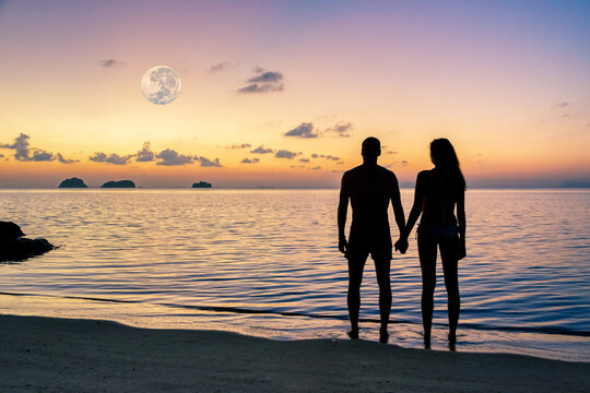 Silhouettes of a young couple standing on a sandy beach holding hands and watching spectacular sunset over the sea and islands and huge rising full moon.