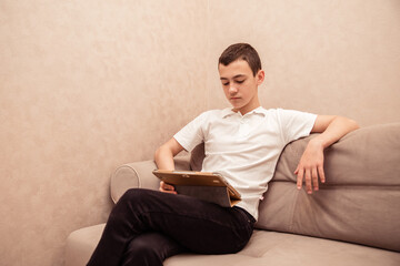 Teenager boy in white T-shirt sits on sofa with laptop