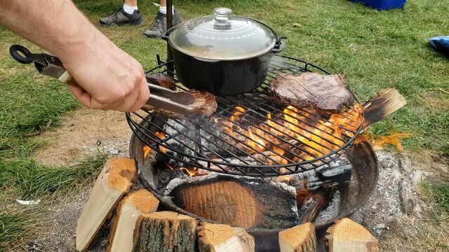 Grilling beef steaks over fire pit next to cast iron pot of boiling soup while at camp site.