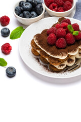 portion of Classic tiramisu dessert with raspberries and blueberries on ceramic plate isolated on white