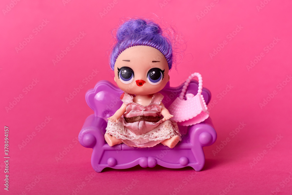 Wall mural Tiny doll with purple hair and big eyes, modern kids toy with rosy dress on pink, doll with big eyes sitting on small sofa against rosy background.