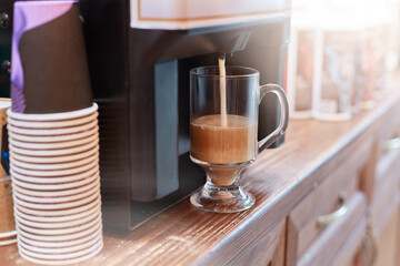 Coffee machine brewing cuppuccino coffee, stack of disposable paper cups, glass of tasty hot beverage on cafe's counter.