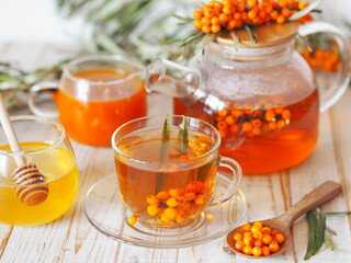 Vitamin healthy sea buckthorn tea in a glass cup with a teapot, honey, jam, fresh raw sea buckthorn berries and sprigs on a white kitchen table. Sea buckthorn berry picking season.