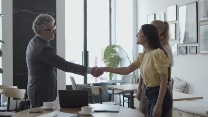 Female customers meeting with mature male legal or financial expert in co-working space, shaking hands over table with cups and smiling. Business communication or handshake concept