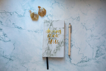 white marble notebook with text dreams come true  and beige pen and yellow earrings on white marble background. Stationery and accessories flatlay.