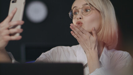 Cheerful woman sending air kiss for mobile selfie in night office