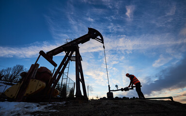 Silhouette of petroleum operator in work vest and helmet using oil pumping unit to extract crude...