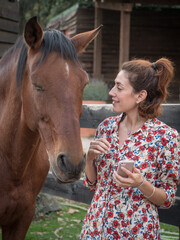 Female with phone in her hand smiling to a chestnut horse.